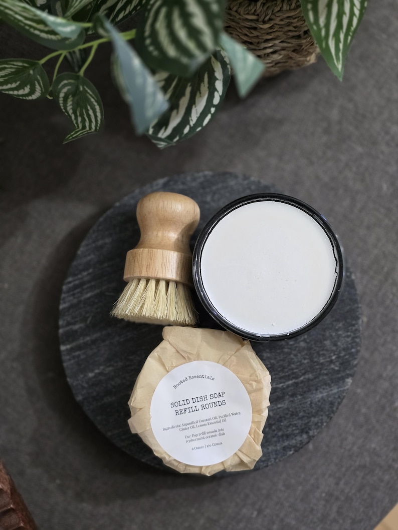 以下が含まれることがあります： 白い丸い食器用石鹸のリフィルバーで、ラベルに「Rooted Essentials Solid Dish Soap Refill Rounds」と書いてあります。石鹸は黒いスレートの表面に置かれ、木製のブラシと白いセラミックの容器があります。