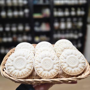 May include: A wicker basket holds several round, off-white soaps, each intricately carved with a sunflower design. The soaps are arranged neatly in the basket, which is being held by a person. The background is a blurred view of shelves with various bottles.