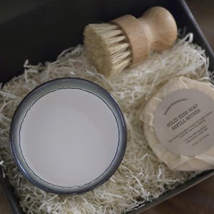 K&ouml;nnte beinhalten: Eine Geschenkbox gef&uuml;llt mit wei&szlig;em Seidenpapier, die eine kleine wei&szlig;e Keramikschale, eine B&uuml;rste aus Holz mit Naturborsten und eine runde Seifenstange enth&auml;lt, die in wei&szlig;es Papier eingewickelt ist, auf dem der Text "Solid Dish Soap Refill Rounds" gedruckt ist.