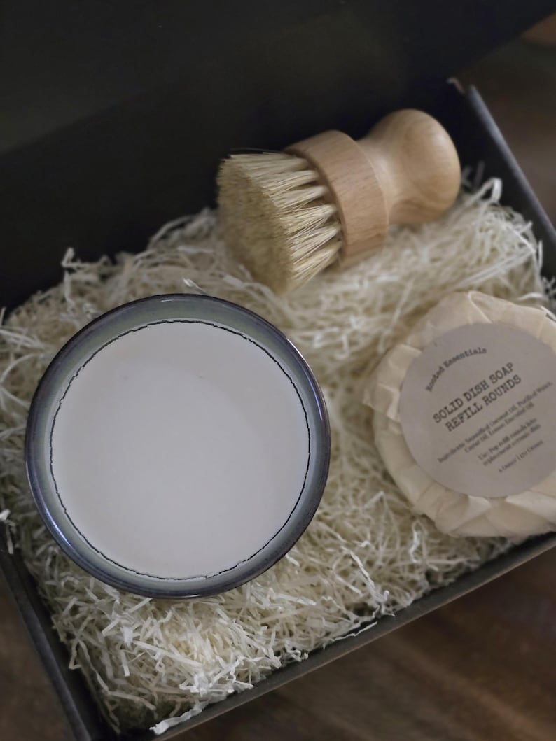 K&ouml;nnte beinhalten: Eine Geschenkbox gef&uuml;llt mit wei&szlig;em Seidenpapier, die eine kleine wei&szlig;e Keramikschale, eine B&uuml;rste aus Holz mit Naturborsten und eine runde Seifenstange enth&auml;lt, die in wei&szlig;es Papier eingewickelt ist, auf dem der Text "Solid Dish Soap Refill Rounds" gedruckt ist.