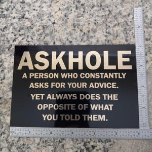 May include: Black sign with gold lettering that reads "ASKHOLE A PERSON WHO CONSTANTLY ASKS FOR YOUR ADVICE. YET ALWAYS DOES THE OPPOSITE OF WHAT YOU TOLD THEM."