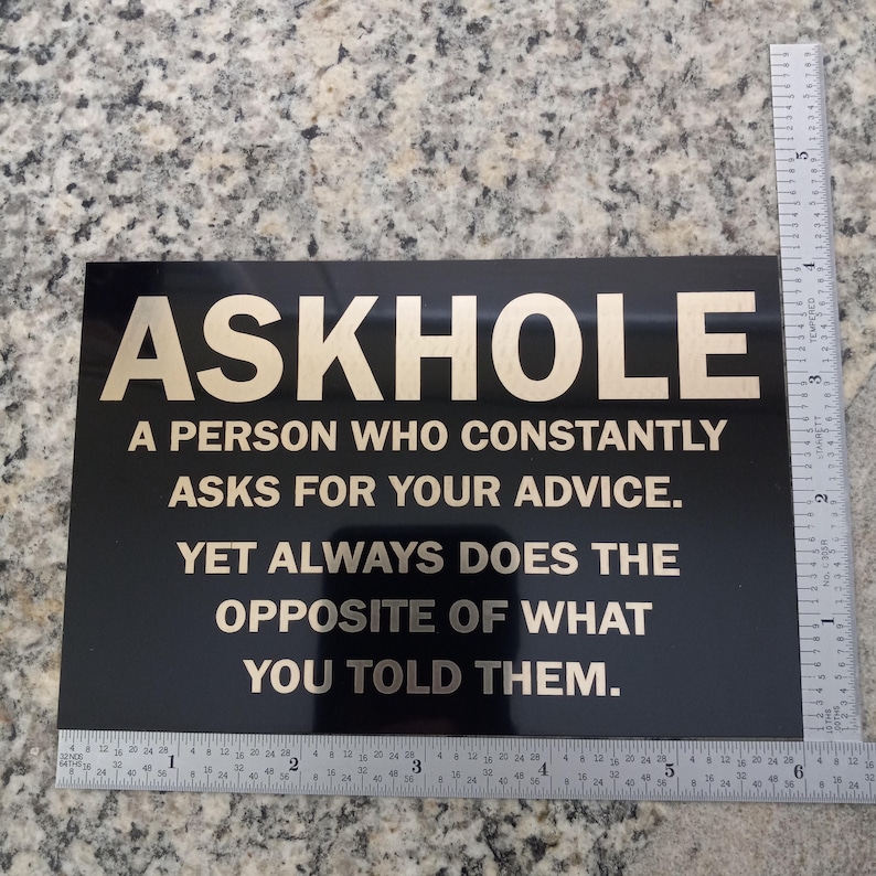 May include: Black sign with gold lettering that reads "ASKHOLE A PERSON WHO CONSTANTLY ASKS FOR YOUR ADVICE. YET ALWAYS DOES THE OPPOSITE OF WHAT YOU TOLD THEM."