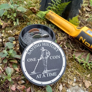 May include: A black and white round sign with the text "Saving History One Coin At A Time" and an illustration of a person using a metal detector. A black container with coins is next to the sign, and a yellow metal detector is in the background.