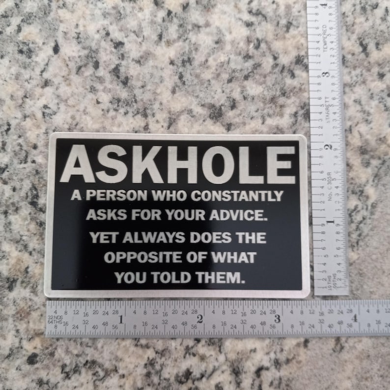 May include: A black and silver sticker with the text "ASKHOLE" in large silver letters. The text below reads "A person who constantly asks for your advice. Yet always does the opposite of what you told them."