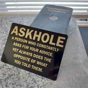 May include: A black sign with gold lettering that reads "ASKHOLE A PERSON WHO CONSTANTLY ASKS FOR YOUR ADVICE. YET ALWAYS DOES THE OPPOSITE OF WHAT YOU TOLD THEM."