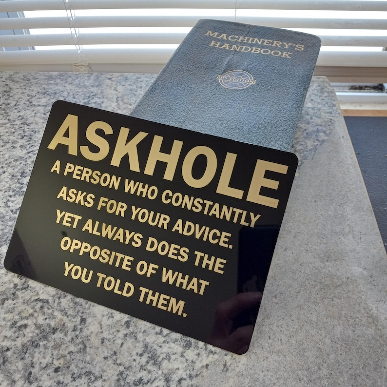May include: A black sign with gold lettering that reads "ASKHOLE A PERSON WHO CONSTANTLY ASKS FOR YOUR ADVICE. YET ALWAYS DOES THE OPPOSITE OF WHAT YOU TOLD THEM."