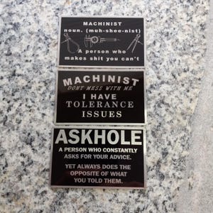 May include: Three black rectangular metal signs with white text. The top sign reads "MACHINIST" and defines the term. The middle sign says "MACHINIST DON'T MESS WITH ME I HAVE TOLERANCE ISSUES." The bottom sign reads "ASKHOLE" and defines the term.