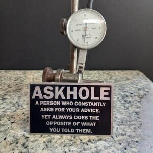 May include: A black and silver metal sign with the text "ASKHOLE" and the definition: "A person who constantly asks for your advice. Yet always does the opposite of what you told them."