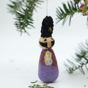 May include: A purple and brown felt ornament shaped like a person wearing a black hat with a yellow star. The ornament has a white face with a black moustache and a yellow smile.