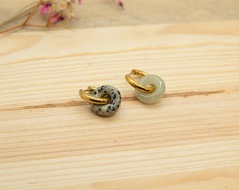 May include: Two pairs of hoop earrings. One pair has a speckled grey stone, the other a pale green stone. Both have gold-coloured hoops. The earrings are displayed on a light wood surface, with a few pink flowers in the background.