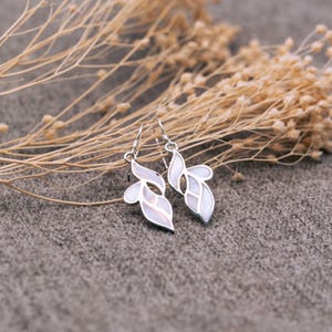 Könnte beinhalten: Ein Paar silberne Ohrringe mit weißen, blattartigen Anhängern. Die Ohrringe sind zart und feminin.