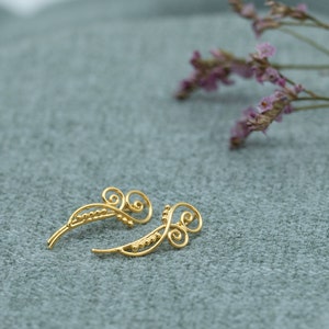 Könnte beinhalten: Ein Paar goldene Ohrclips mit einem zarten, filigranen Design. Die Ohrclips sind aus dünnem Draht gefertigt und haben kleine Perlen entlang der Länge.