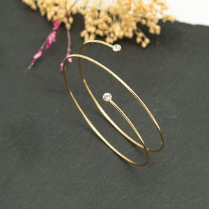 May include: Two gold-toned arm cuffs with a minimalist design. Each cuff features a delicate, curved band with a sparkling, round, clear crystal accent. The jewelry is displayed on a dark gray surface, with dried floral accents in the background.