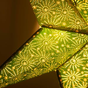 Puede incluir: Primer plano de una linterna de papel en forma de estrella verde con un patrón detallado de pequeñas estrellas y puntos. La linterna está iluminada desde dentro, creando un brillo cálido y acogedor. El diseño es simétrico e intrincado.
