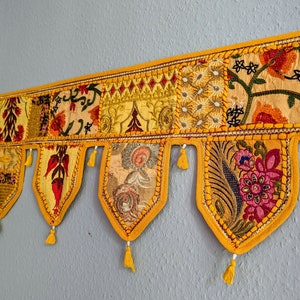 Op de afbeelding: Een decoratieve, rechthoekige wandhanger met een gele rand en meerdere stoffen panelen. De panelen hebben verschillende patronen, waaronder bloemen en geometrische ontwerpen, in tinten geel, oranje en rood. Elk paneel heeft een decoratieve kwast. De wandhanger is ongeveer 91 cm breed.