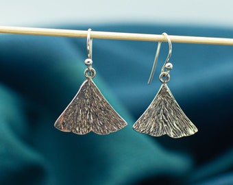 Puede incluir: Un par de pendientes de plata con forma de hojas de ginkgo. Los pendientes cuelgan de una varilla de madera sobre un fondo azul.