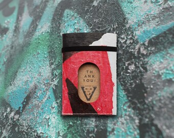 Può includere: Un piccolo taccuino nero, rosso e bianco con un cerchio ritagliato che rivela una carta marrone con il testo "Thank You!" e un piccolo grafico di una scimmia. Il taccuino ha un elastico nero.