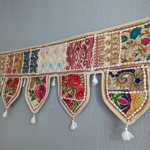 Könnte beinhalten: Ein dekorativer Wandbehang mit Patchwork-Design. Er zeigt verschiedene bestickte Stoffquadrate in Rot-, Gold-, Blau- und Cremetönen mit Blumen- und Paisley-Mustern. Weiße Quasten hängen von unten, und goldfarbene Zierleisten akzentuieren die Kanten.