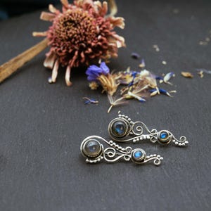 Puede incluir: Un par de pendientes de plata con un diseño delicado y en espiral. Cada pendiente presenta tres piedras redondas de labradorita, una más grande y dos más pequeñas.