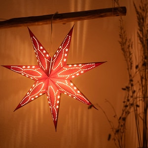 May include: A red paper star-shaped lantern with white star-shaped cutouts, suspended from a wooden beam. The lantern is illuminated, creating a warm glow and shadows. A decorative item for the home.