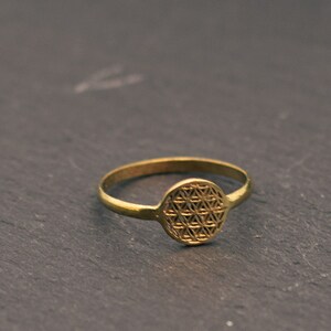 Op de afbeelding: Een goudkleurige ring met een cirkelvormig, geometrisch ontwerp. De ring heeft een smalle band en een rond middelpunt met een gedetailleerd, symmetrisch patroon. De ring ligt op een donkere, getextureerde ondergrond.