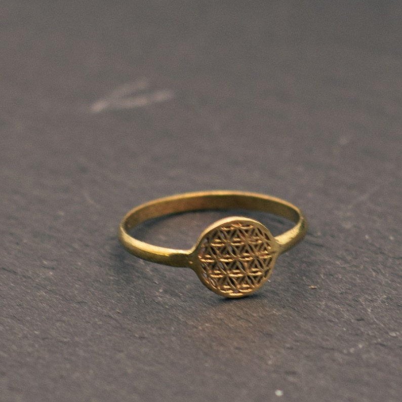 Op de afbeelding: Een goudkleurige ring met een cirkelvormig, geometrisch ontwerp. De ring heeft een smalle band en een rond middelpunt met een gedetailleerd, symmetrisch patroon. De ring ligt op een donkere, getextureerde ondergrond.