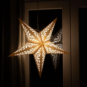 May include: A decorative, illuminated star-shaped paper lantern. The star has five points and is adorned with a repeating, white, floral-like pattern. The warm light shines through the cutouts, creating a cozy ambiance. The star is hanging in front of a window.