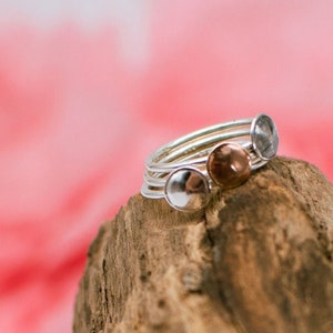 Könnte beinhalten: Ein Satz von fünf Silberringen mit gehämmerter Textur. Drei der Ringe haben runde, glatte Oberseiten, während zwei eine leicht gewölbte Form haben. Die Ringe sind auf einem Stück Treibholz gestapelt.