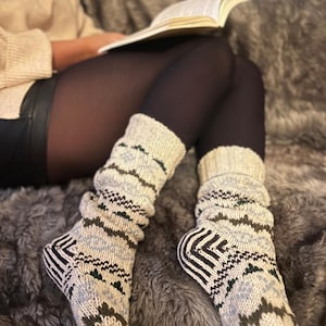 May include: A pair of cozy, cream-colored knitted socks with a geometric pattern in shades of blue, green, and brown. The socks are worn by a person sitting on a fuzzy, gray rug.