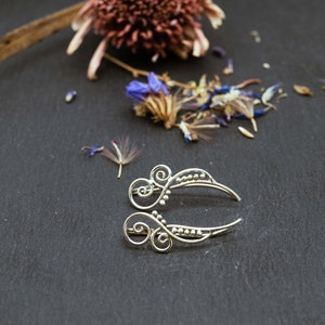 Könnte beinhalten: Ein silberner Ohrclip mit einem filigranen, komplizierten Design mit kleinen Perlen und Spiralen. Der Clip ist gebogen und hat eine Halbmondform.