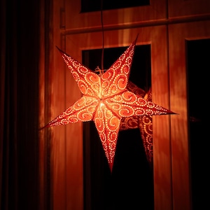 Op de afbeelding: Een vijfhoekige, stervormige hanglamp van rood papier met ingewikkelde uitgesneden krul- en stippatronen. De lamp wordt van binnenuit verlicht en werpt een warme rode gloed door de perforaties heen op de omgeving.
