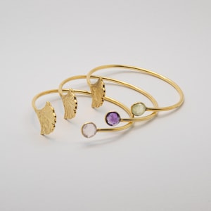 Può includere: Braccialetti in metallo dorato. Due braccialetti presentano dettagli a forma di ala, mentre gli altri hanno pietre preziose rotonde rosa, viola e verdi. I braccialetti sono aperti e sembrano delicati.