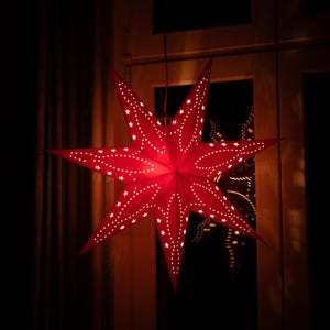 May include: A seven-pointed star-shaped paper lantern in a vibrant red hue. The surface features intricate, laser-cut star patterns and linear perforations that allow light to shine through, creating a glowing effect against the dark surroundings.