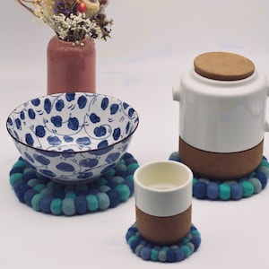 Könnte beinhalten: Ein Set aus drei Untersetzern aus blauem und grünem Filz mit einer weißen Keramikschüssel, einer weißen Keramiktasse und einer weißen Keramikteekanne mit einem Korkdeckel. Schüssel und Tasse haben Korkböden.