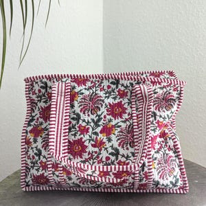 Pode incluir: Uma sacola tote acolchoada com um padrão floral em tons de vermelho, amarelo e verde sobre um fundo branco. A sacola tem acabamento e alças listradas em vermelho e branco. Está sobre uma superfície redonda e cinza.