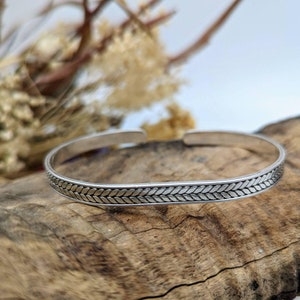 May include: A silver bangle bracelet with a braided design. The bracelet is on a wooden surface.