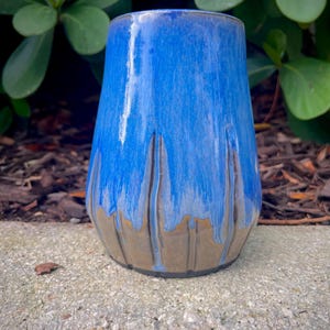 May include: A cylindrical ceramic tumbler featuring a vibrant blue glaze on the upper portion that transitions into a smooth, earthy brown base. The blue glaze drips downward in vertical streaks, creating a textured effect over the brown surface.