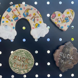 May include: A collection of handmade ceramic art pieces with the word "FUCK" and "FUCK THIS" on them. The pieces include a rainbow, a heart, a circle, and a multi-sided shape. The rainbow and heart have colorful patterns.