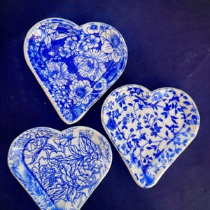 May include: Three heart-shaped ceramic dishes with blue and white floral patterns. Each dish features a different design, including detailed flower and leaf motifs. The dishes are set against a dark blue background.