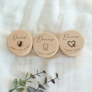 Könnte beinhalten: Drei runde Holzkisten mit eingraviertem Text und Symbolen. Die Kisten sind mit "Dana's Umbilical Cord", "Dana's First Tooth" und "Dana's First Hair" beschriftet.