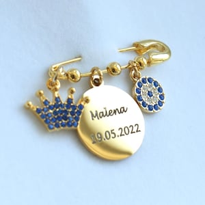 Könnte beinhalten: Goldene Sicherheitsnadel mit einer blauen Krone, einem runden Anhänger mit dem Namen Malena und dem eingravierten Datum 19.05.2022 sowie einem blauen Glücksbringer-Anhänger.