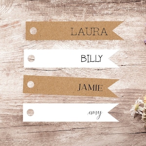 May include: Four brown and white paper place cards with the names Laura, Billy, Jamie, and Amy written on them. A bouquet of wildflowers in shades of purple, yellow, and white is on a wooden surface.
