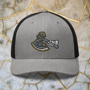 Puede incluir: Una gorra de camionero gris y negra con un diseño de hacha bordado en blanco y negro. El hacha tiene un contorno amarillo y un mango blanco.