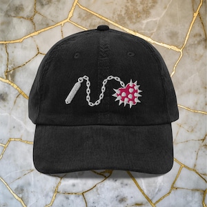 Puede incluir: Gorra de béisbol de pana negra con un diseño bordado. El diseño presenta un corazón rosa con púas blancas, conectado por una cadena a un objeto cilíndrico blanco. La gorra tiene una visera curva y un botón superior.