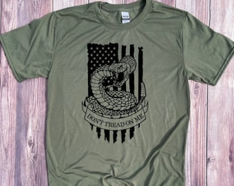 May include: A green t-shirt featuring a black and white image of a rattlesnake coiled around an American flag. The text "Don't Tread On Me" is written on a banner below the snake.