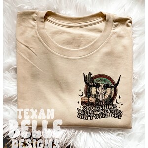 May include: Tan t-shirt with a skull and crossbones graphic, cactus, and two glasses of whiskey. The text reads "Something's tellin' me this ain't over yet".