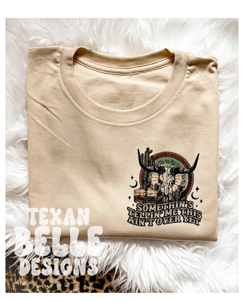 May include: Tan t-shirt with a skull and crossbones graphic, cactus, and two glasses of whiskey. The text reads "Something's tellin' me this ain't over yet".