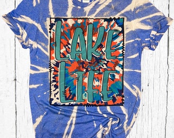 Puede incluir: Una camiseta azul tie-dye con un diseño de blanqueador blanco. La camiseta tiene un gráfico cuadrado con el texto "Lake Life" en letras azul verdoso y rojas.