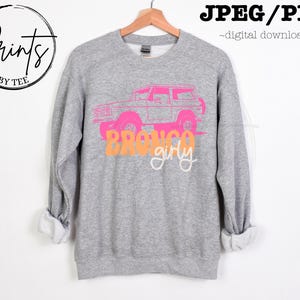 May include: Gray sweatshirt with a pink and orange graphic of a vintage Bronco truck with the text "Bronco girly".