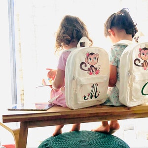 Könnte beinhalten: Zwei weiße Canvas-Rucksäcke mit Affen-Illustrationen und personalisiert mit den Buchstaben "M" und "C". Die Rucksäcke werden von zwei Kindern getragen, die auf einer Holzbank sitzen.